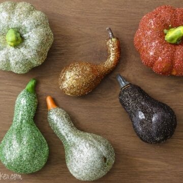 Collection of glitter-covered pumpkins and gourds in green, orange, gold, and black displayed on a wooden surface for DIY fall decor inspiration.