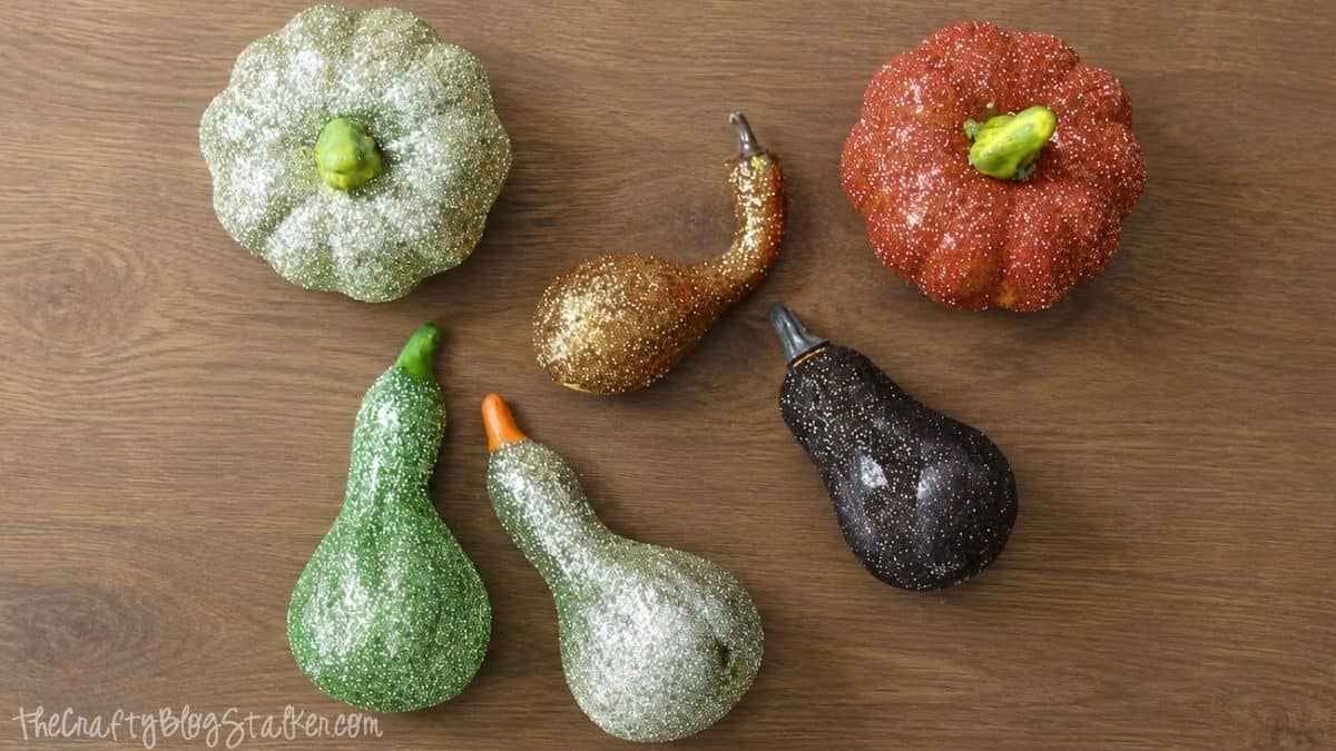 Collection of glitter-covered pumpkins and gourds in green, orange, gold, and black displayed on a wooden surface for DIY fall decor inspiration.