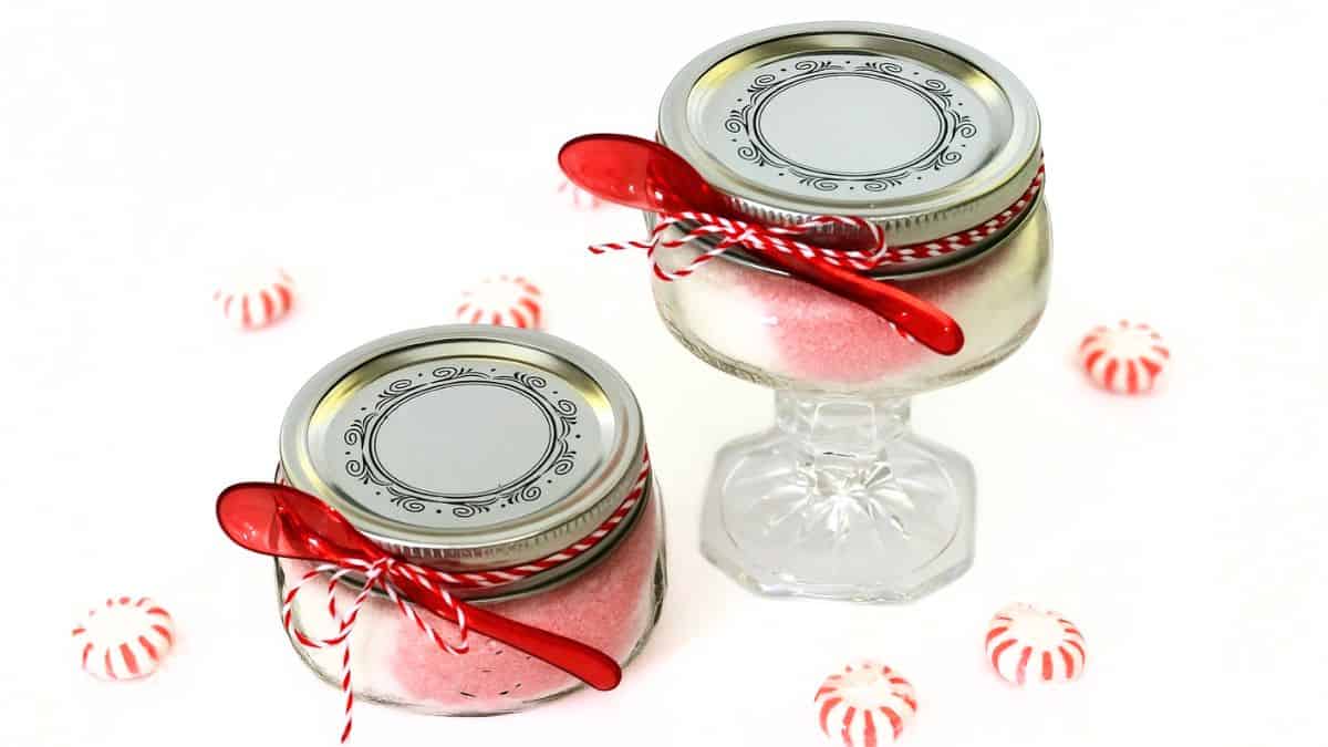 Two jars of homemade peppermint sugar scrub with red and white twine and red spoons, surrounded by peppermint candies on a white background.