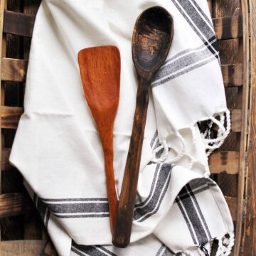Two hand-carved wooden utensils, a dark spoon and a lighter spatula, displayed on a white kitchen towel with black stripes.