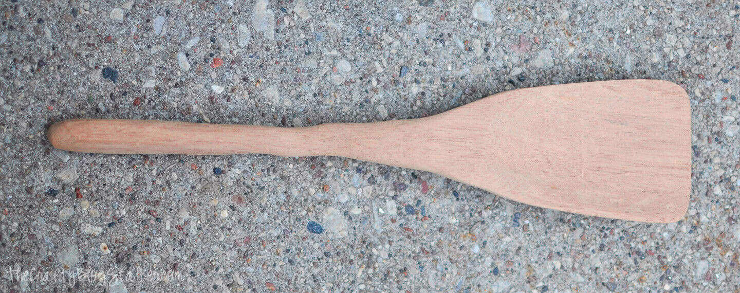 A freshly carved wooden spatula lying on concrete, showing its smooth handle and wide, sanded paddle-shaped end.