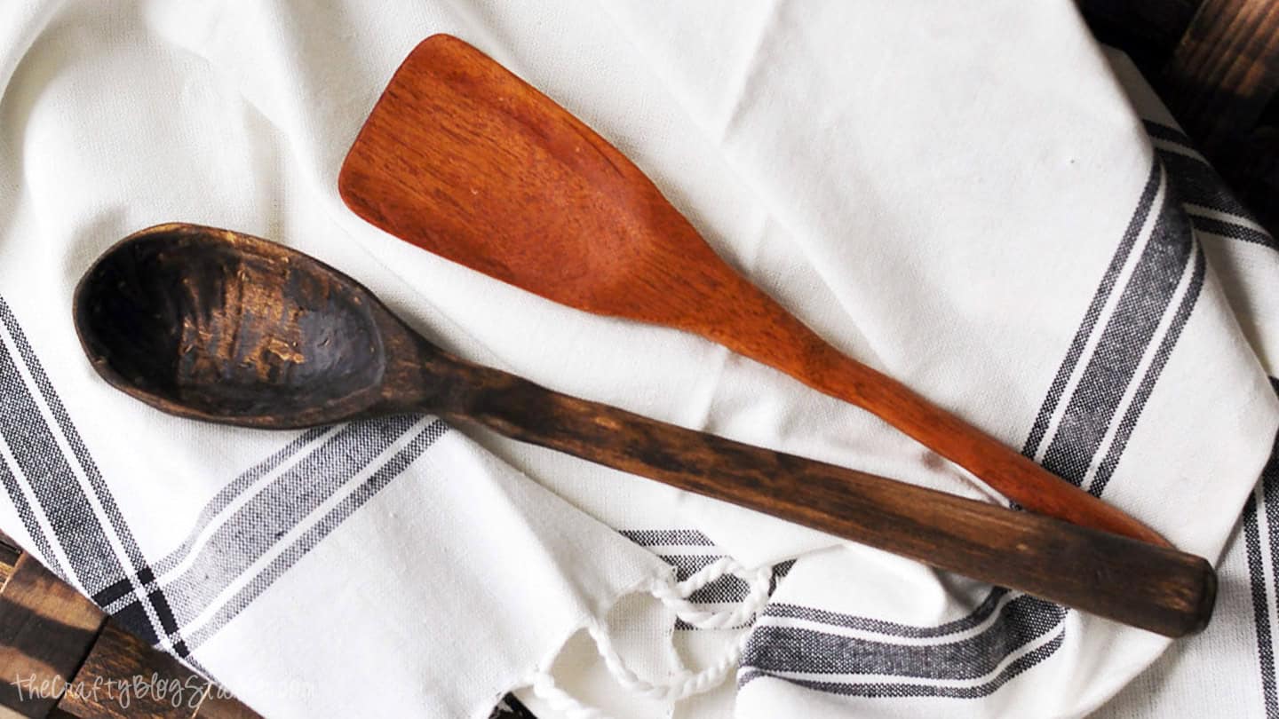 Two hand-carved wooden kitchen utensils, a dark spoon and a lighter spatula, resting on a white dish towel with black stripes.