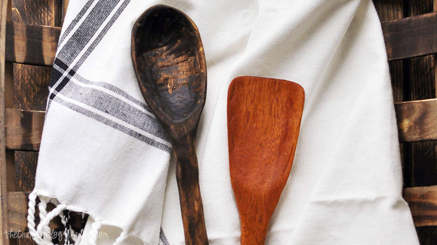 Two hand-carved wooden utensils, a dark spoon and a lighter spatula, displayed on a white striped kitchen towel.