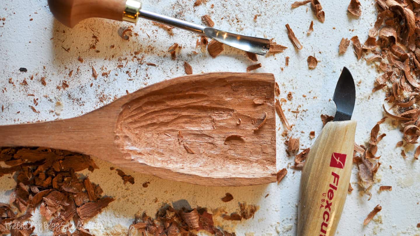 A wooden spoon in progress with the bowl being carved, surrounded by wood shavings and carving tools on a work surface.