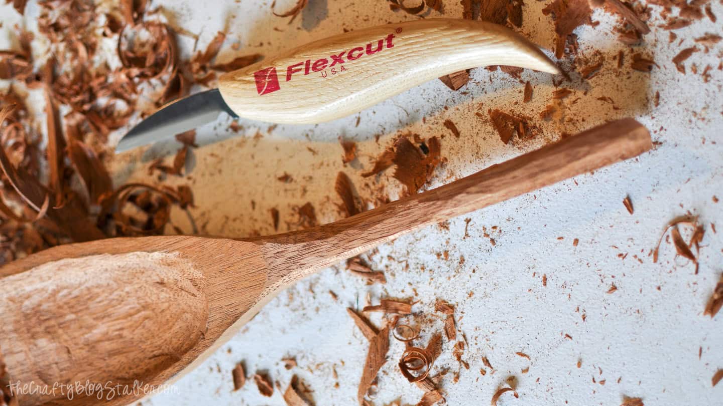 A partially carved wooden spoon lying on a work surface covered in wood shavings, with a Flexcut carving knife nearby.