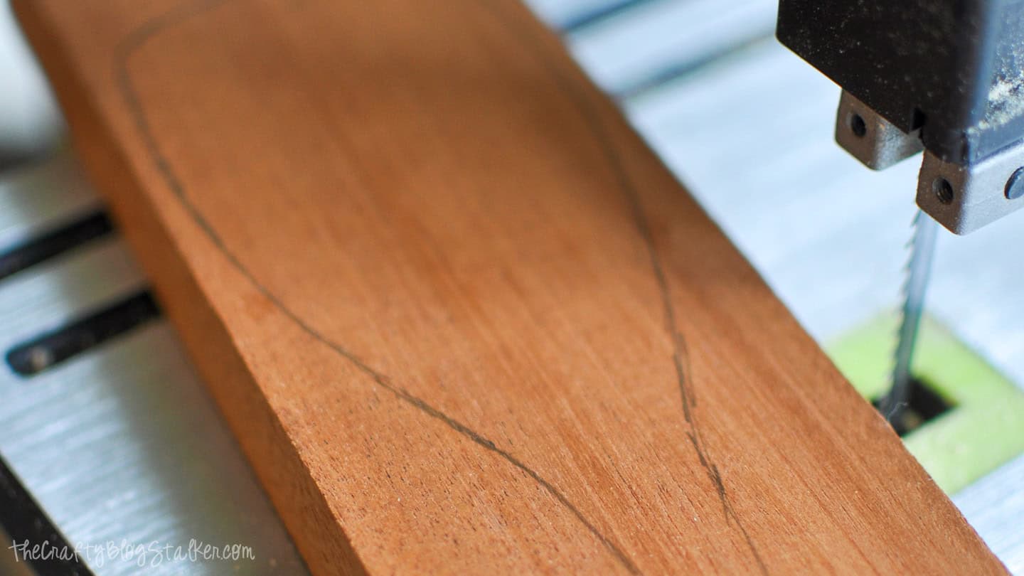 Close-up of a wooden board with a spoon outline being cut on a bandsaw, showing the blade and part of the saw table.