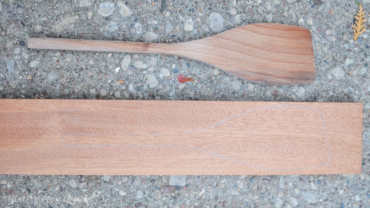 Wooden spoon blank with a spoon shape sketched on a board, shown next to a partially carved wooden spoon on a concrete surface.