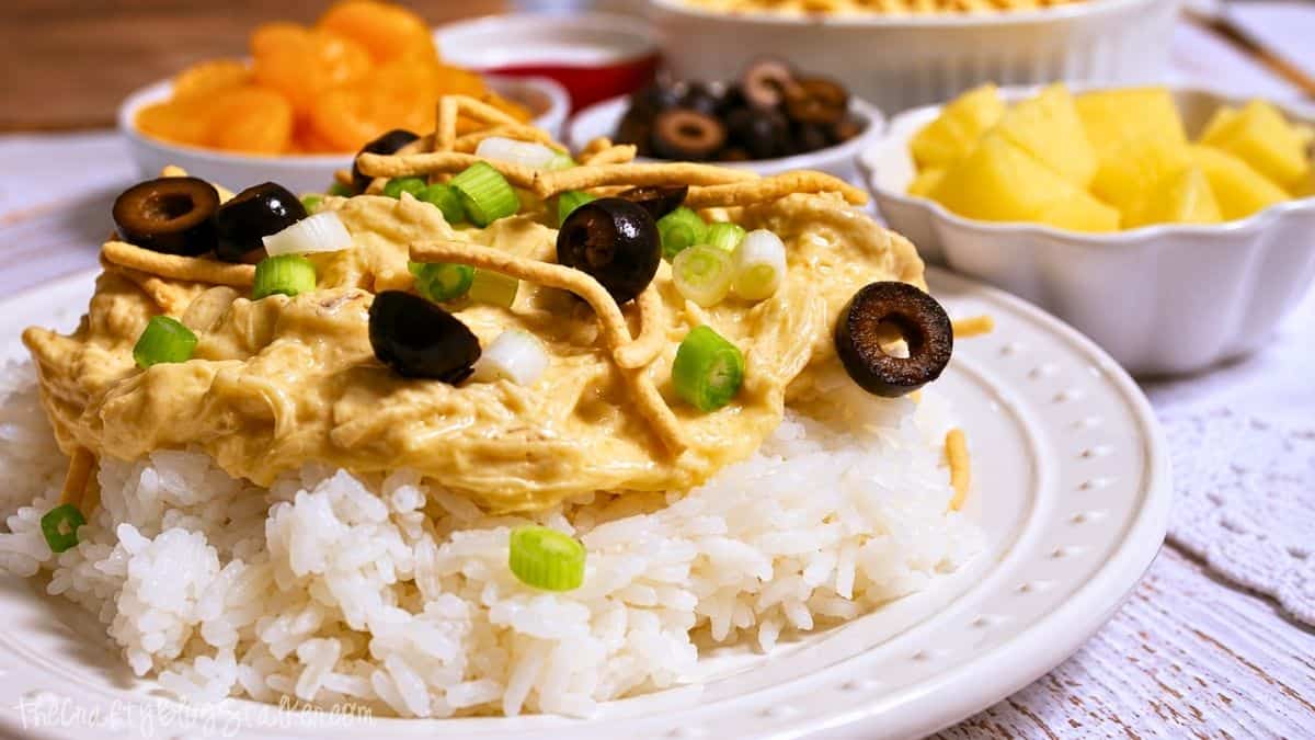 Plate of Hawaiian haystacks with creamy chicken over rice, topped with chow mein noodles, black olives, and green onions, with fruit toppings in the background.