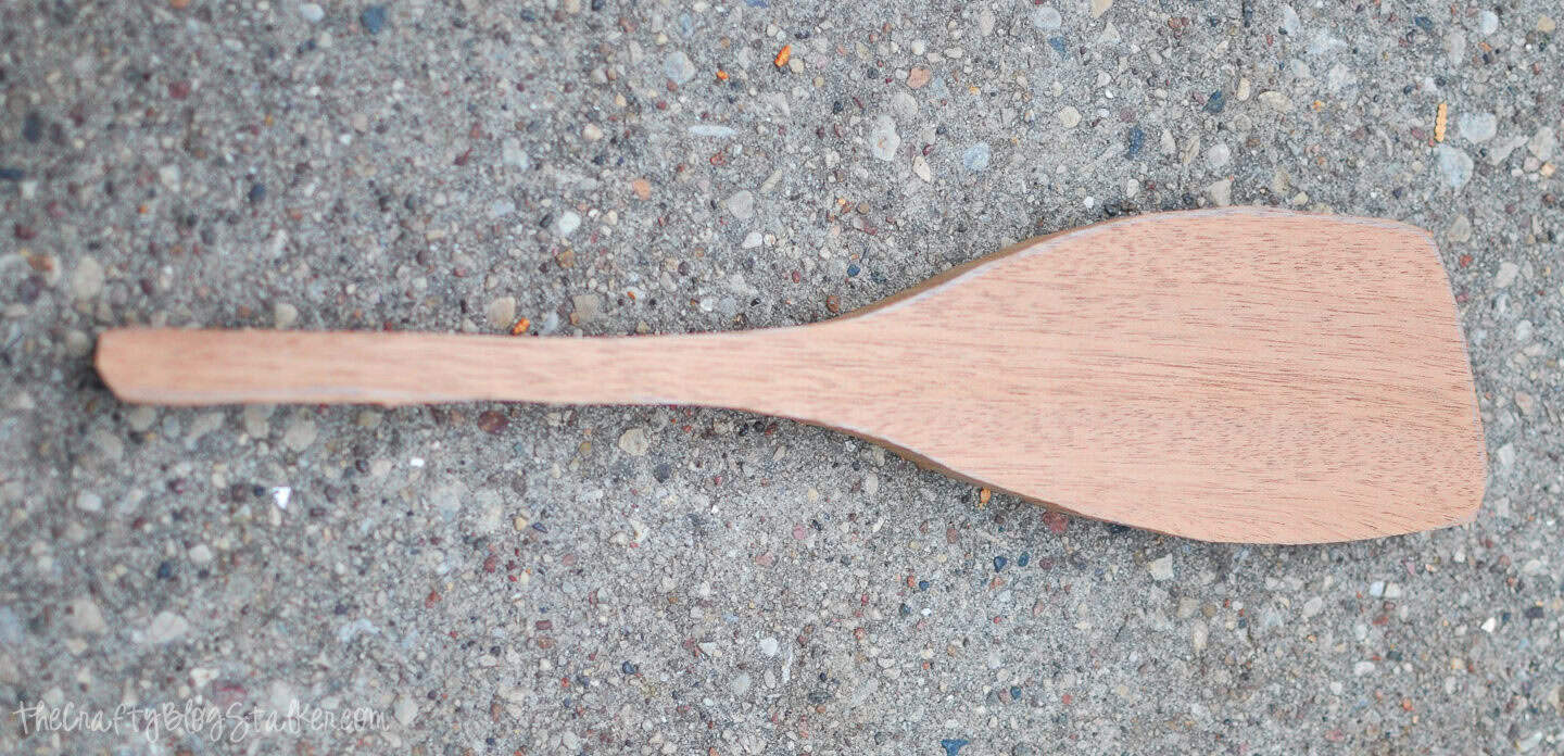 A carved wooden spoon blank with a shaped handle and flat paddle end resting on a rough concrete surface.