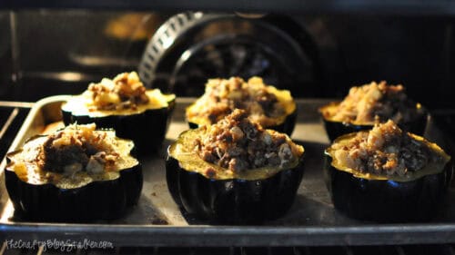 Stuffed acorn squash halves filled with sausage and apple stuffing baking on a sheet pan inside the oven until golden and heated through.