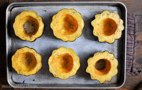 Roasted acorn squash halves seasoned with salt and pepper on a baking sheet, ready to be filled with sausage and apple stuffing.