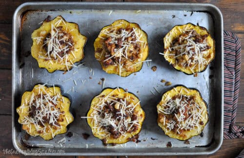 Stuffed acorn squash halves filled with sausage and apples, topped with Parmesan cheese, and arranged on a baking sheet before baking.