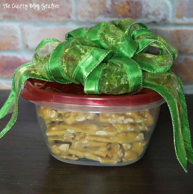 Peanut Brittle in a reusable plastic container with a green bow on top.