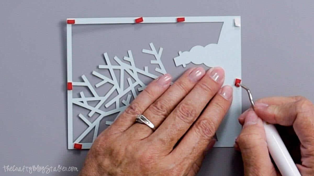 Hands use a weeding tool to place red foam adhesive squares on the back of a light blue cardstock layer for a Christmas card.
