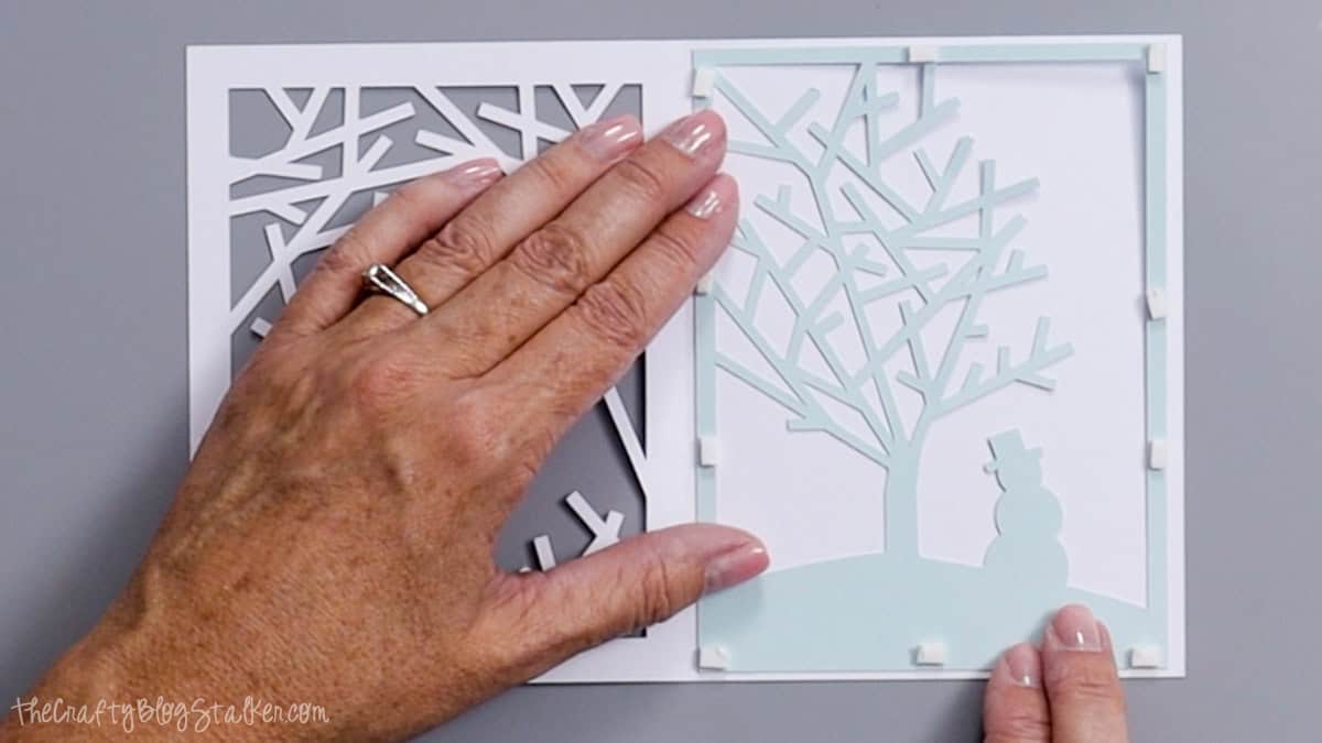 Hands position a light blue cardstock layer with a tree and snowman onto a white Christmas card using foam adhesive squares.