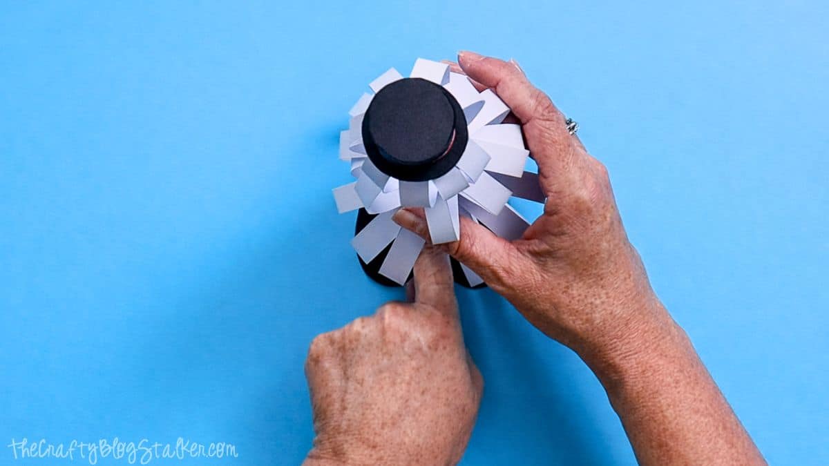 Hands attaching paper strip snowman feet to the bottom of a finished paper strip snowman, viewed from above on a blue work surface.