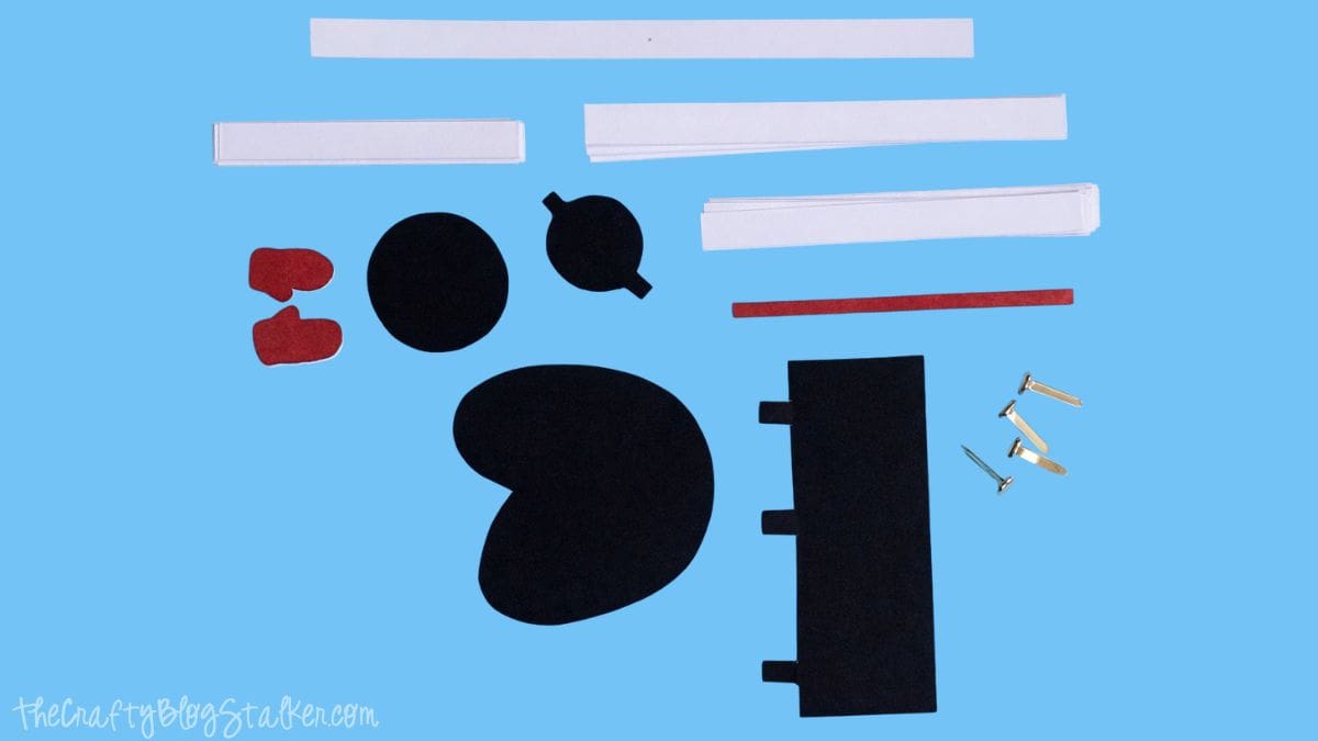 All cut paper pieces for a paper strip snowman craft laid out, including strips, body shapes, hat pieces, mittens, and brads on a work surface.