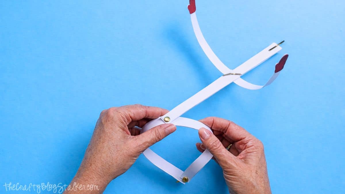 Hands bending connected paper strips with metal brads to form a rounded snowman body, with attached arms and mitten cutouts on a blue background.