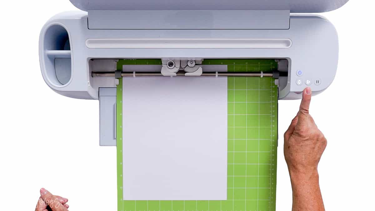 White cardstock on a green Cricut StandardGrip cutting mat loaded into a Cricut machine as a hand presses the start button.