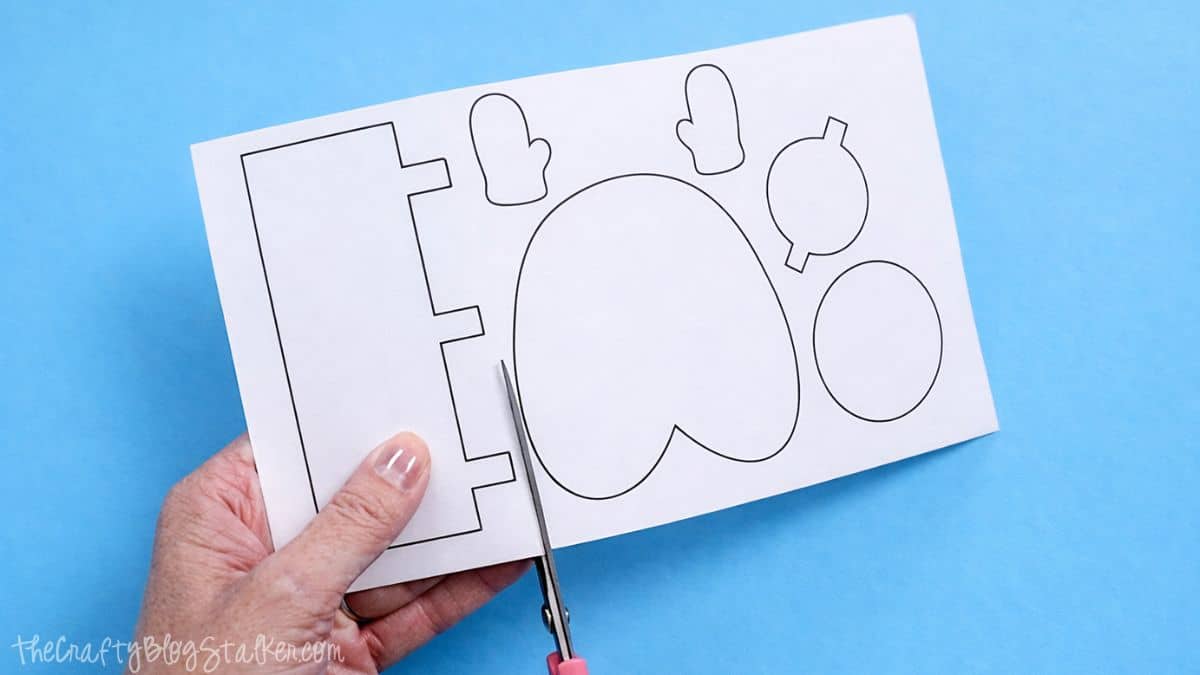Hand cutting printed snowman template shapes from cardstock with scissors on a craft table during a paper strip snowman project.