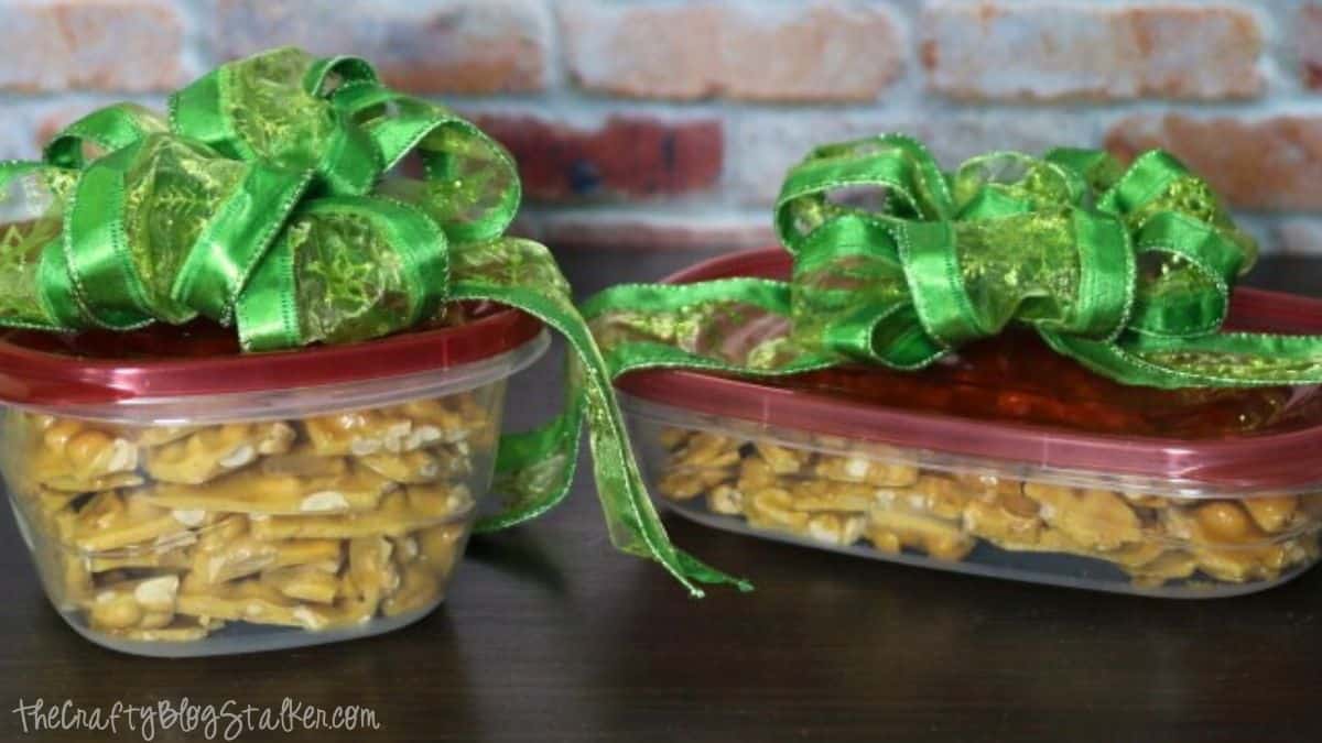 Two plastic containers filled with homemade peanut brittle, each topped with a red lid and decorated with a large green ribbon bow for gifting.