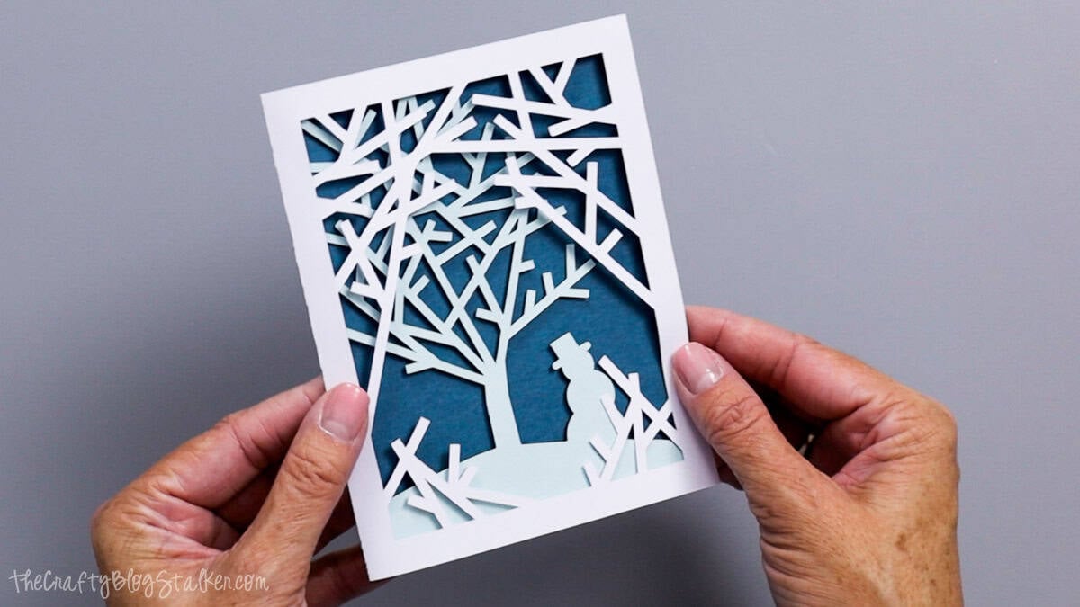 Hands hold a finished layered Christmas card featuring white snowy tree branches, a light blue snowman, and a dark blue background.