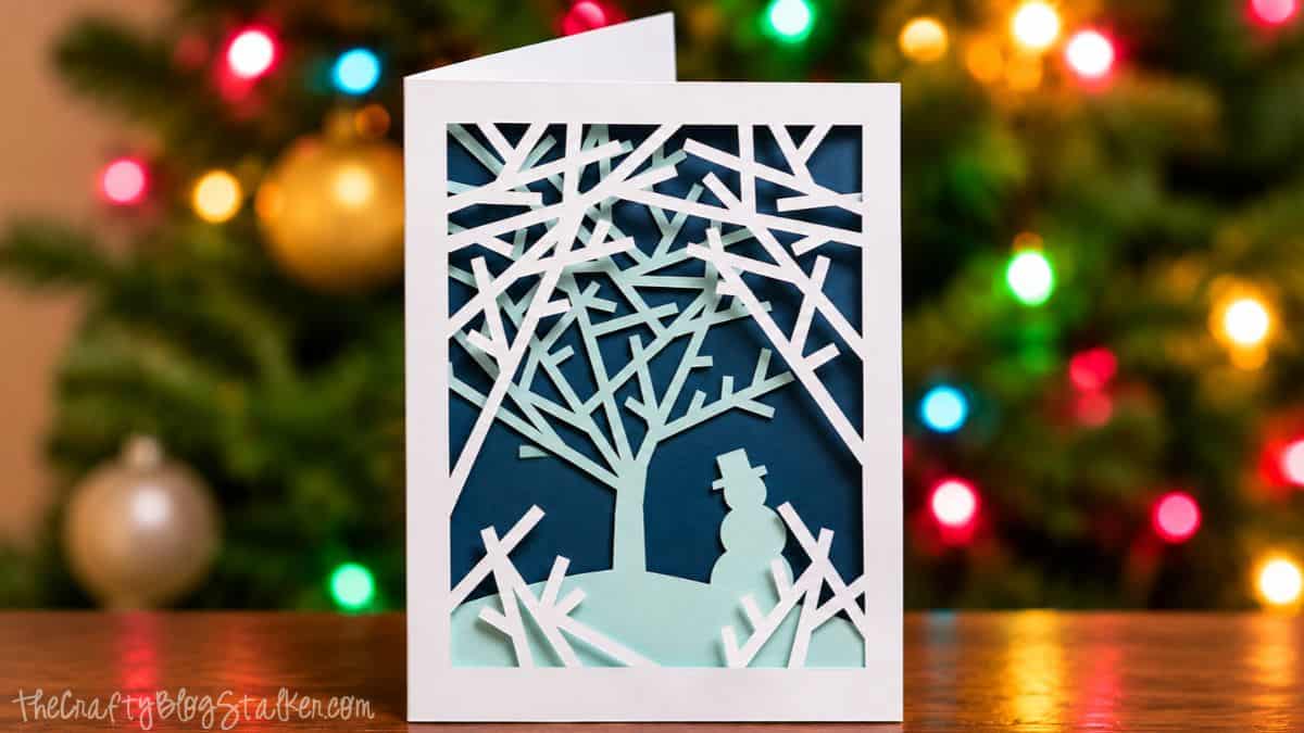 Layered Snowy Woods Christmas card with white paper branches, a mint green tree and snowman, displayed in front of colorful Christmas lights.