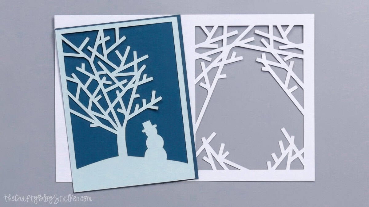 Layered Christmas card pieces cut from white, light blue, and dark blue cardstock, featuring snowy tree branches and a snowman.