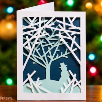 Layered Snowy Woods Christmas card with white paper branches, a mint green tree and snowman, displayed in front of colorful Christmas lights.