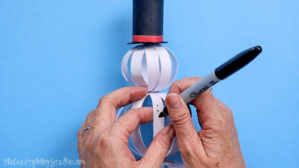 Hands drawing coal buttons on a paper strip snowman body with a marker, while the snowman stands upright wearing a paper top hat.