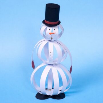 Paper strip snowman craft standing upright, made from layered paper strips with a top hat, carrot nose, and mitten hands on a plain background.