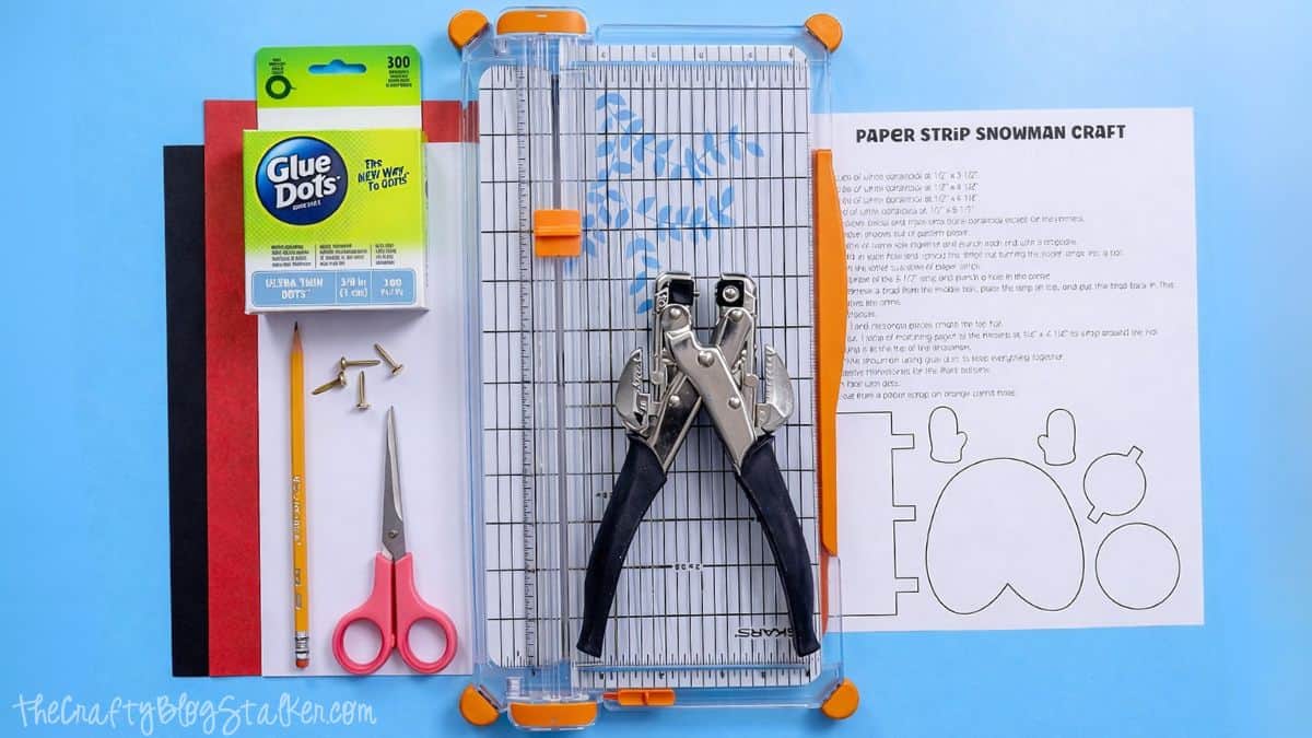 Flat lay of paper strip snowman craft supplies including cardstock sheets, glue dots, pencil, scissors, brads, a paper trimmer, hole punch, and printed instructions.