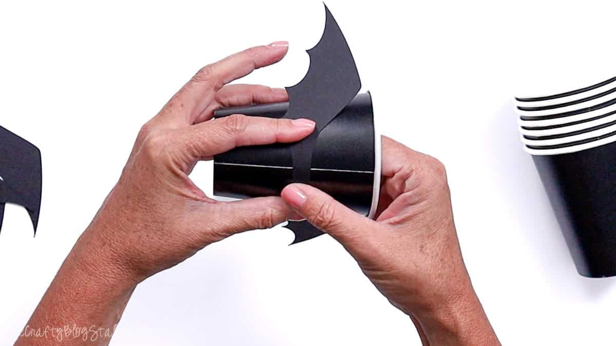Hands attaching black paper bat wings to the side of a black paper cup as part of a Halloween DIY bat treat cup craft project.
