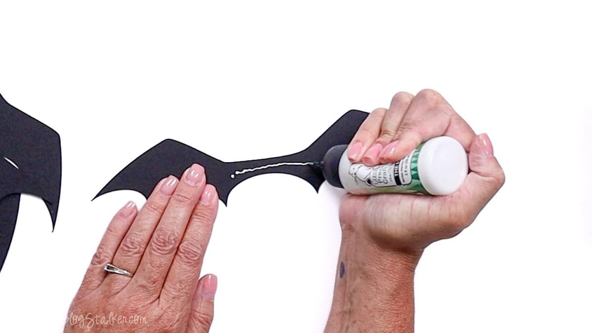 Hands applying craft glue to the center of a black paper bat wing cutout, preparing it for a Halloween DIY project.