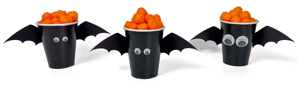 Three black paper cups decorated as bats with wings and googly eyes, each filled with cheese balls for a fun Halloween party treat.