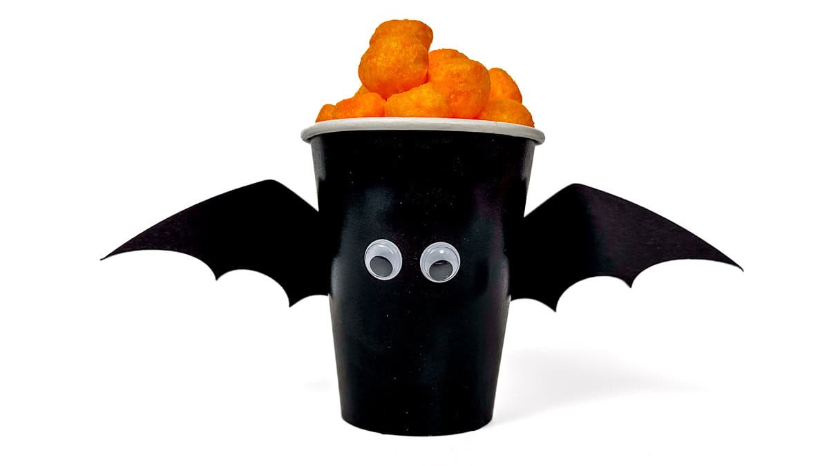 Black paper cup decorated as a bat with googly eyes and paper wings, filled with cheese balls for a fun Halloween treat.