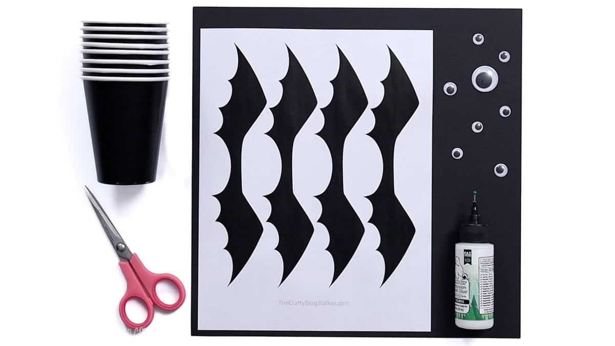 DIY bat treat cup supplies including black cups, scissors, bat wing template, googly eyes, and craft glue laid out on a white background.