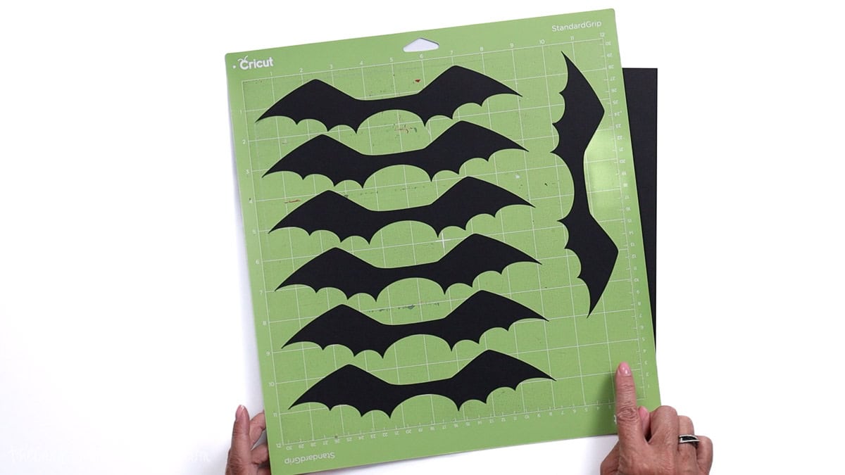 Cricut cutting mat with black cardstock bat wing shapes freshly cut, ready for assembling Halloween craft projects.