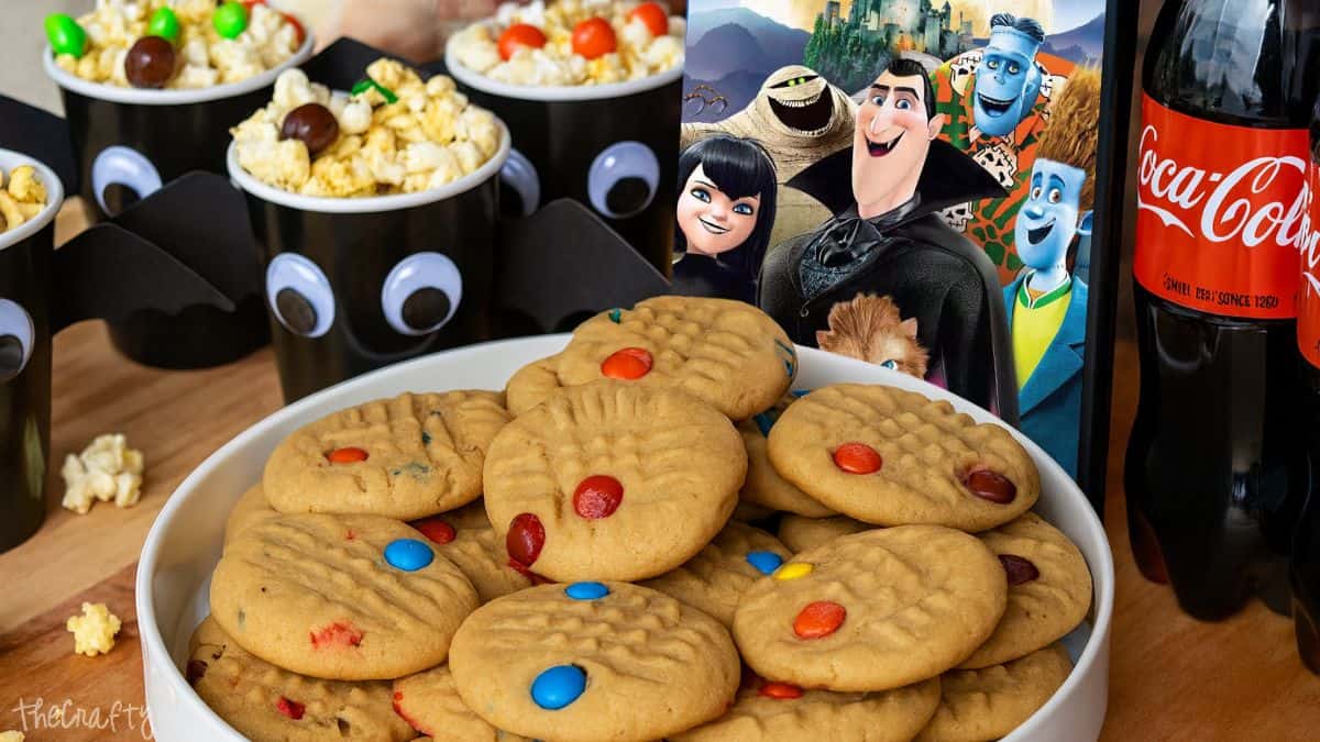 Plate of peanut butter cookies with candy-coated chocolates, bat-themed popcorn cups, Coca-Cola bottles, and a Hotel Transylvania movie poster for a fun movie night.