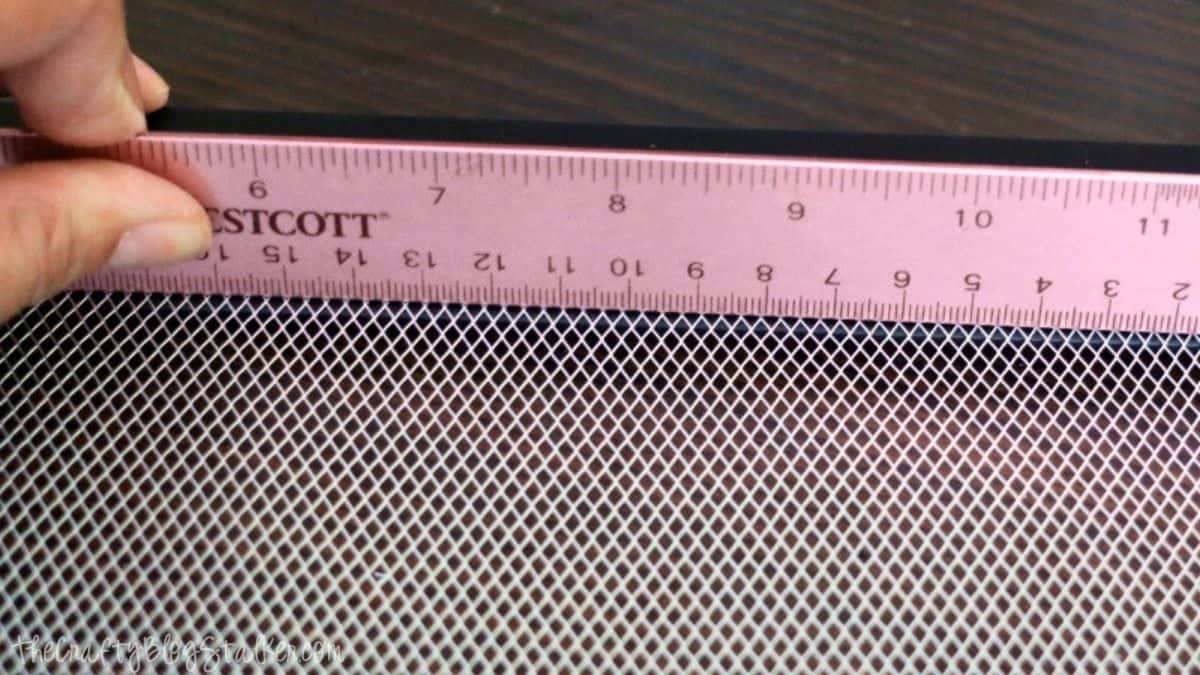 Hand holding a ruler along the edge of a sheet of metal mesh, measuring it on a wooden surface.