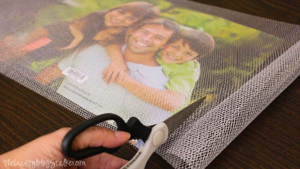 Scissors cutting a piece of metal mesh placed over a photo frame backing on a wooden surface.