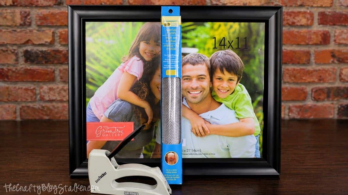 Black 14x11 photo frame, roll of metal mesh, and staple gun displayed as supplies for making a DIY earring holder.
