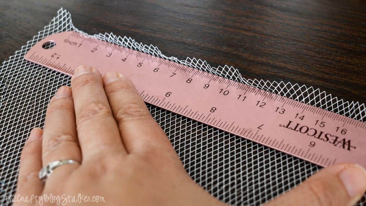 Hand using a ruler to fold and align a piece of metal mesh on a wooden surface.