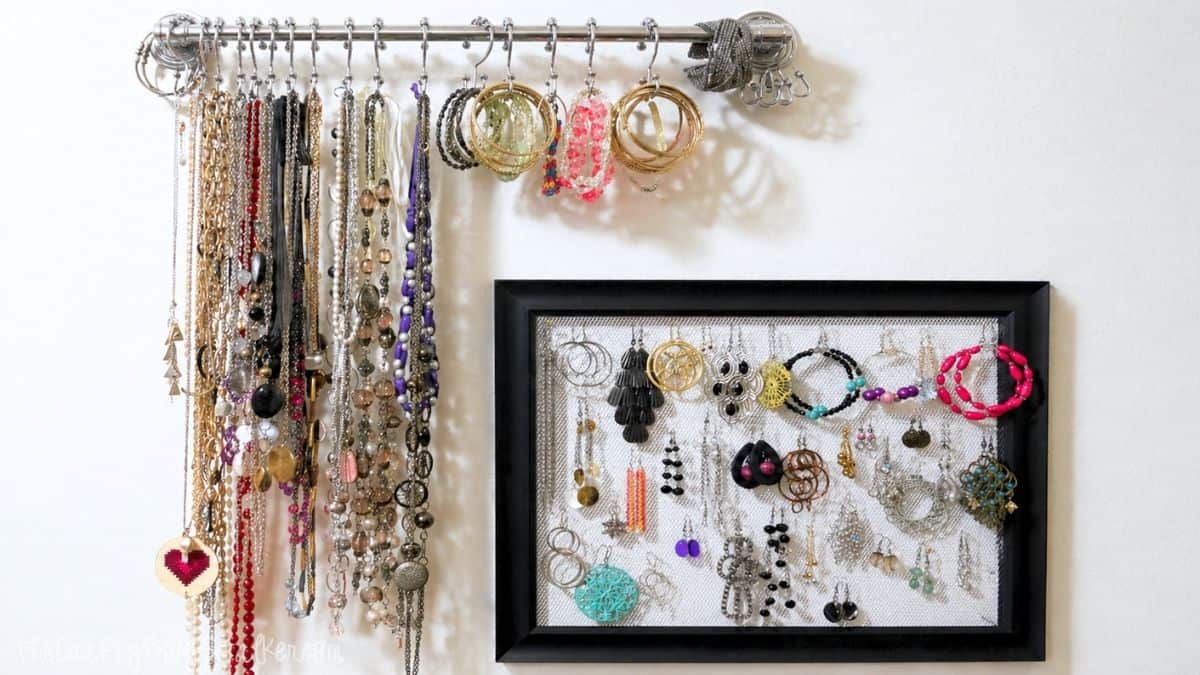 Wall-mounted jewelry organizer with necklaces on a rod and earrings displayed in a black frame with metal mesh backing.