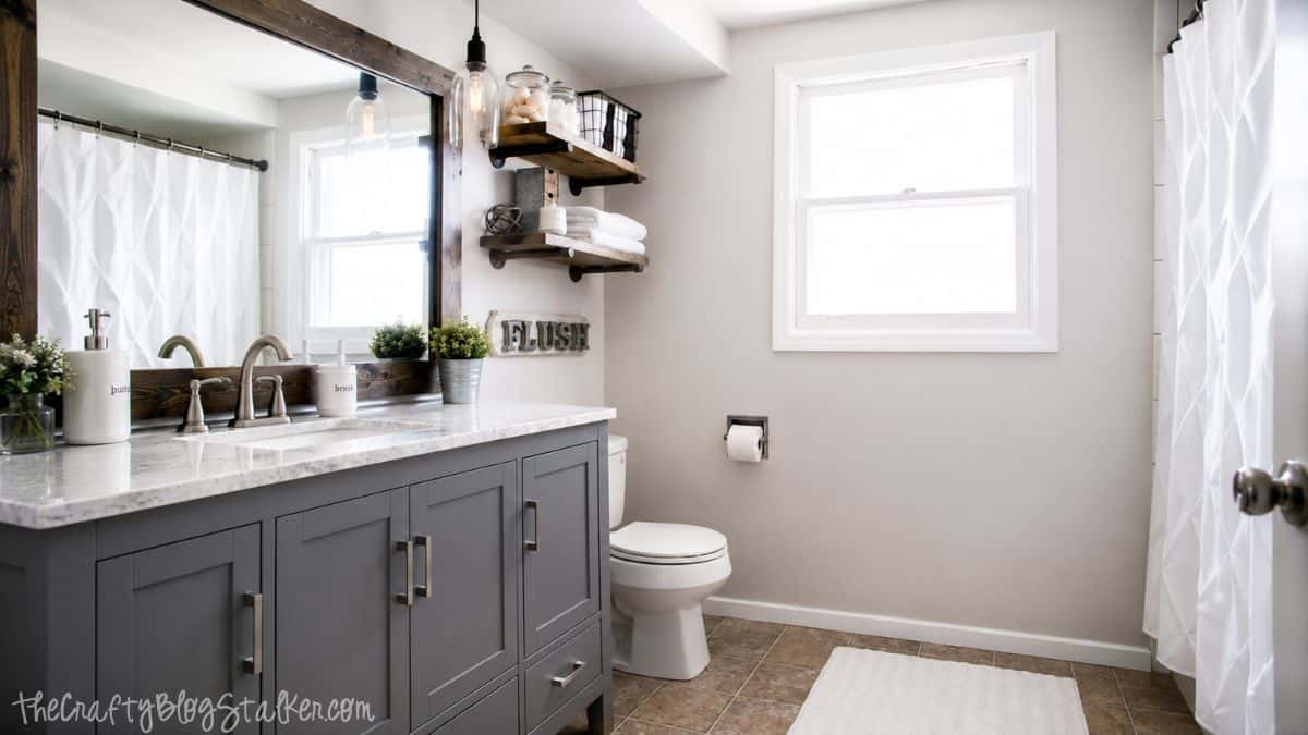 Modern bathroom remodel with gray vanity, marble-look countertop, wood-framed mirror, floating shelves, and white shower curtain.