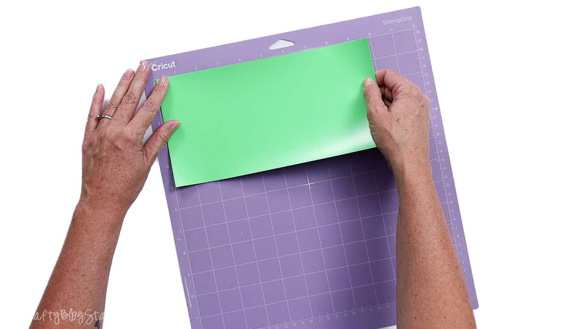 Hands placing a sheet of vinyl onto a purple Cricut StrongGrip cutting mat, smoothing it flat before cutting.