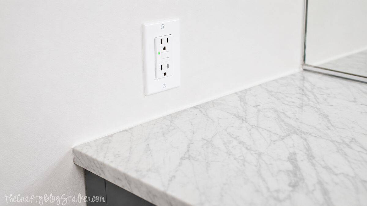 Close-up of a marble-look bathroom vanity countertop with a white wall outlet beside a bathroom mirror.
