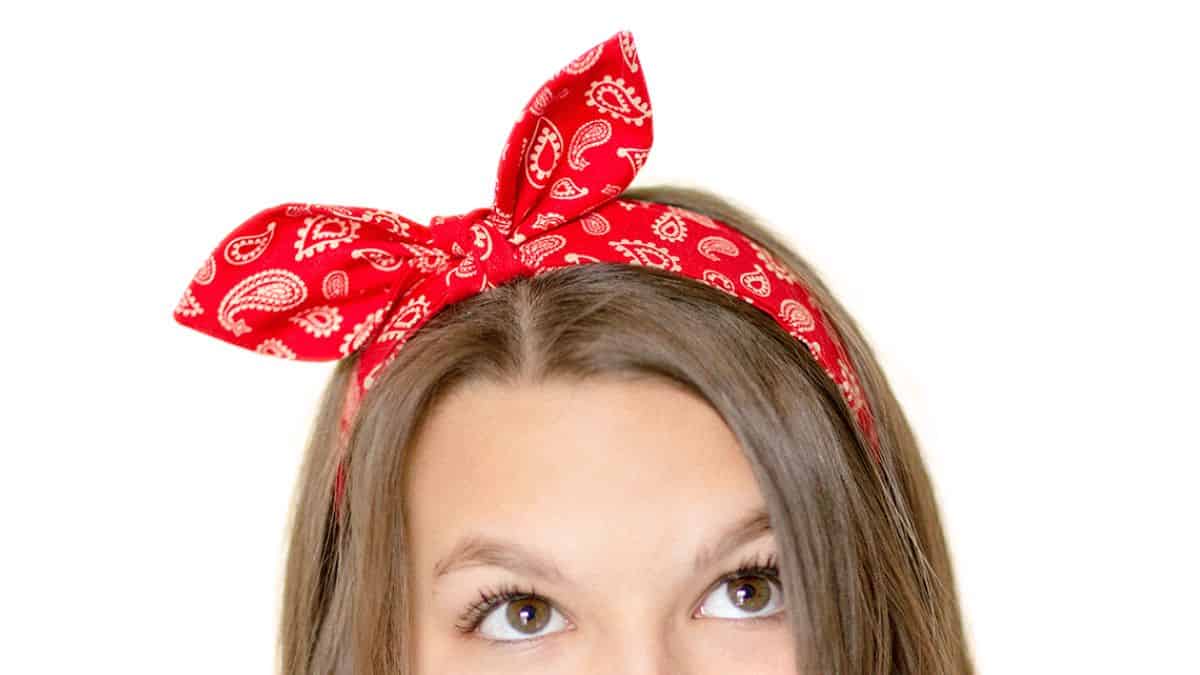 Young girl wearing a red paisley bow tie headband with a large tied bow on top against a white background.