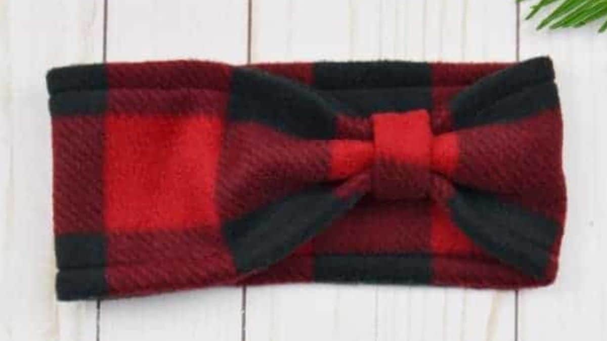Red and black buffalo plaid fleece ear warmer headband with a matching bow displayed on a white wood background.