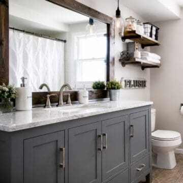 Modern bathroom with gray vanity, marble-look countertop, wood-framed mirror, floating shelves, and brushed nickel faucet.